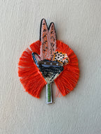 Backside of decorative pin with orange fringe and multicolored design on a light gray background