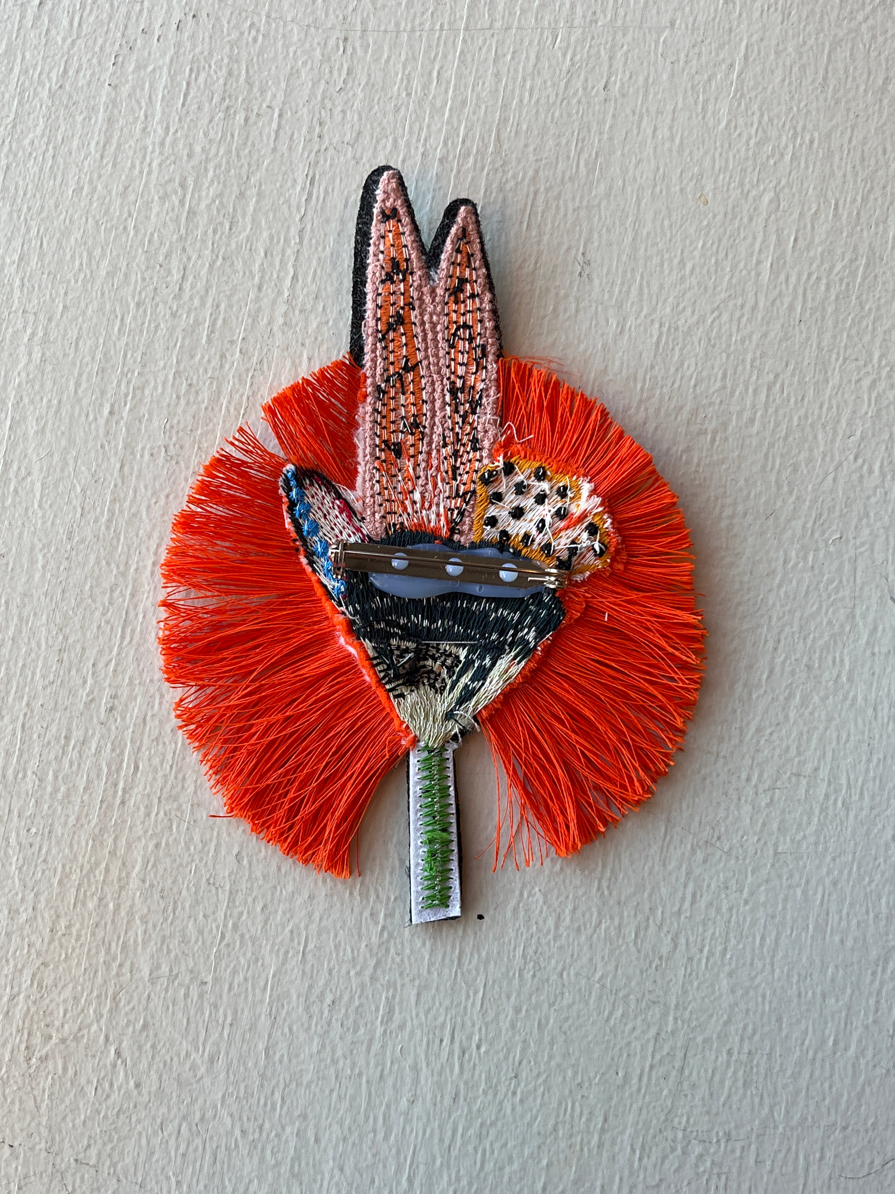 Backside of decorative pin with orange fringe and multicolored design on a light gray background