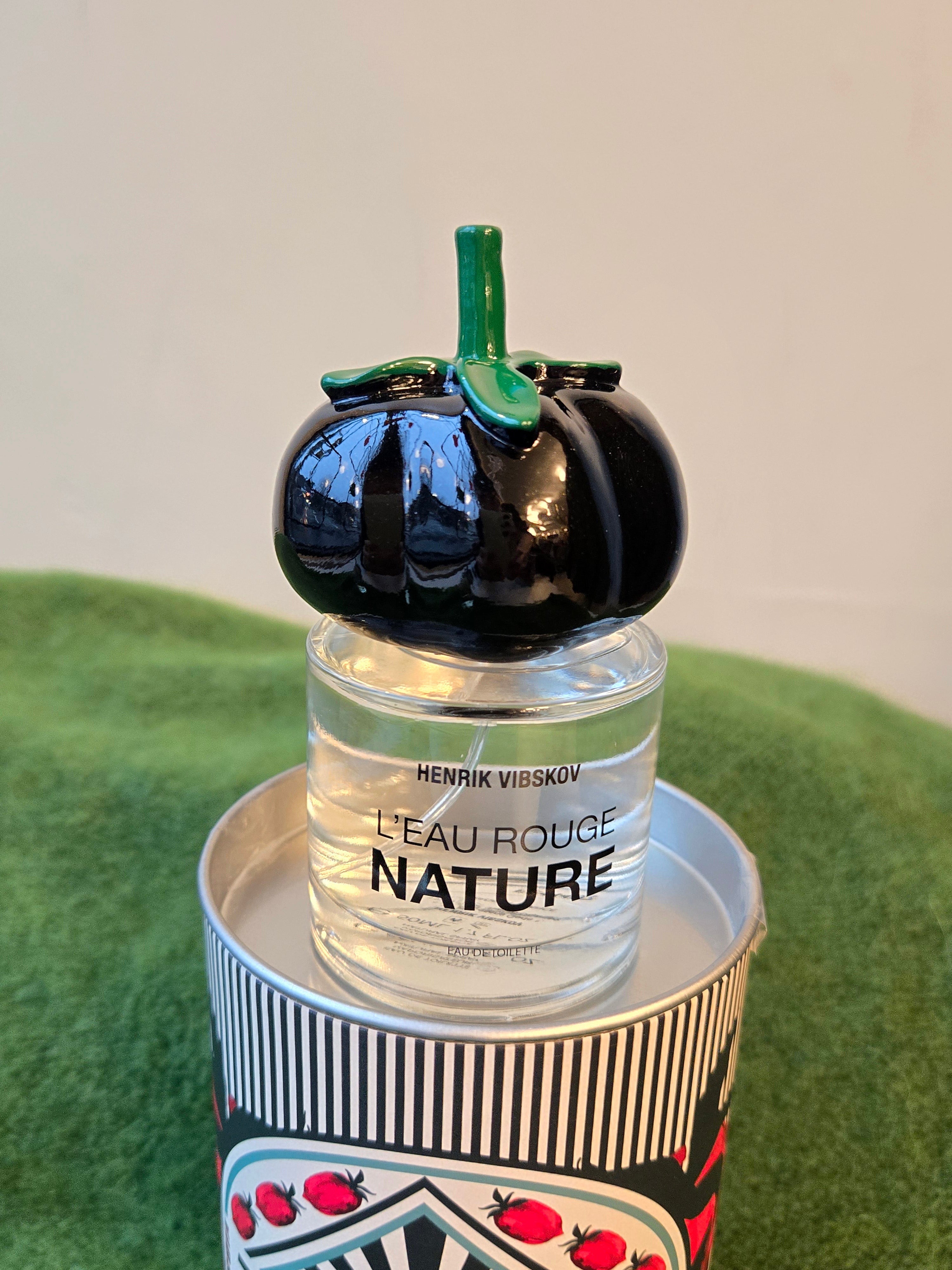 Perfume bottle labeled 'L'Eau Rouge Nature' by Henrik Vibskov with a decorative black tomato cap on a green surface.