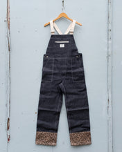 Load image into Gallery viewer, W&#39;menswear Holiday Dungaree features a leopard print fabric at the end of its legs, well contrasted to its overall dark denim fabric and off-white detailing. The brass hardware adds an extra bit of warmth! It has got a front chest pocket that can be zipped up, and two hip pockets in the front.
