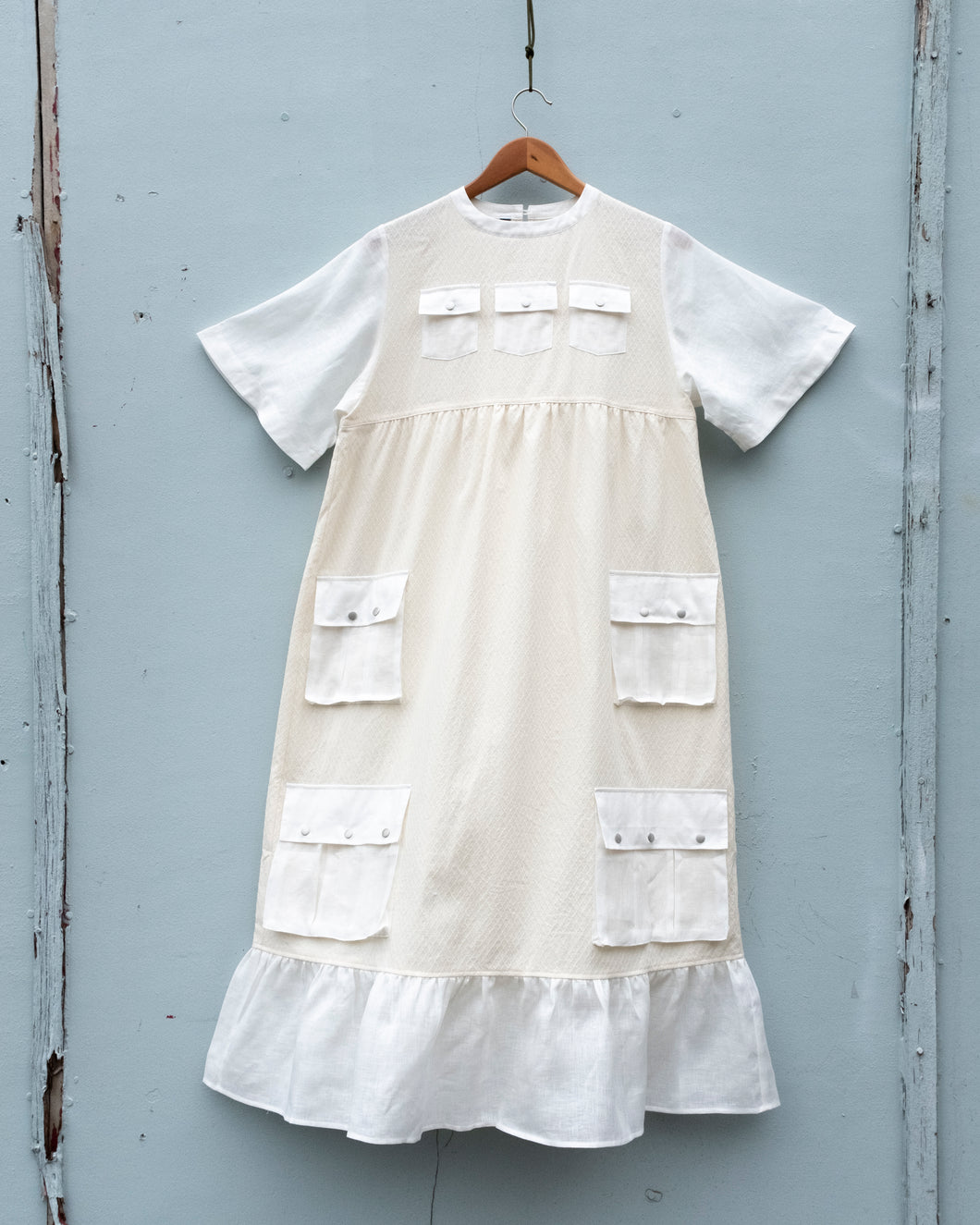 In this image, the W'menswear Summer Fortune Pocket Dress in Off-white is photographed flat. The dress features 3 small chest pockets, who medium sized pockets at hip level, and another pair of large ones below, before the peplum hem at the bottom. All with silver snap buttons.
